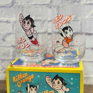 May include: Two clear drinking glasses with cartoon character illustrations. The glasses feature a black-haired character in a red and white outfit. The glasses sit on a yellow box with the words "Ribbon Orange" and "Ribbon Citron" printed on it.