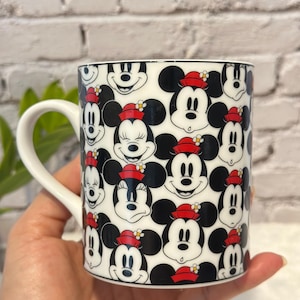 Puede incluir: Taza de cer&aacute;mica blanca con un patr&oacute;n repetido de caras de Mickey Mouse con orejas negras y sombreros rojos. La taza tiene un asa blanca y un borde negro. El dise&ntilde;o incluye a Mickey y Minnie Mouse.