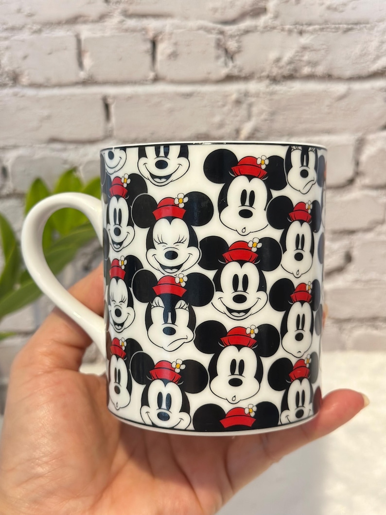 Puede incluir: Taza de cer&aacute;mica blanca con un patr&oacute;n repetido de caras de Mickey Mouse con orejas negras y sombreros rojos. La taza tiene un asa blanca y un borde negro. El dise&ntilde;o incluye a Mickey y Minnie Mouse.
