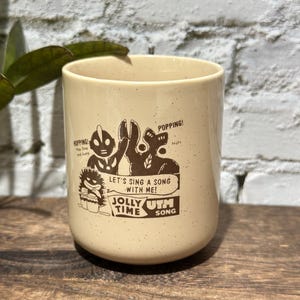 May include: A cream-colored ceramic mug with brown speckles featuring a graphic illustration of characters and the text "HOPPING! Hop Step and Jump", "POPPING! Nuts", "LET'S SING A SONG WITH ME!", "JOLLY TIME", and "UTM SONG" printed in brown ink.