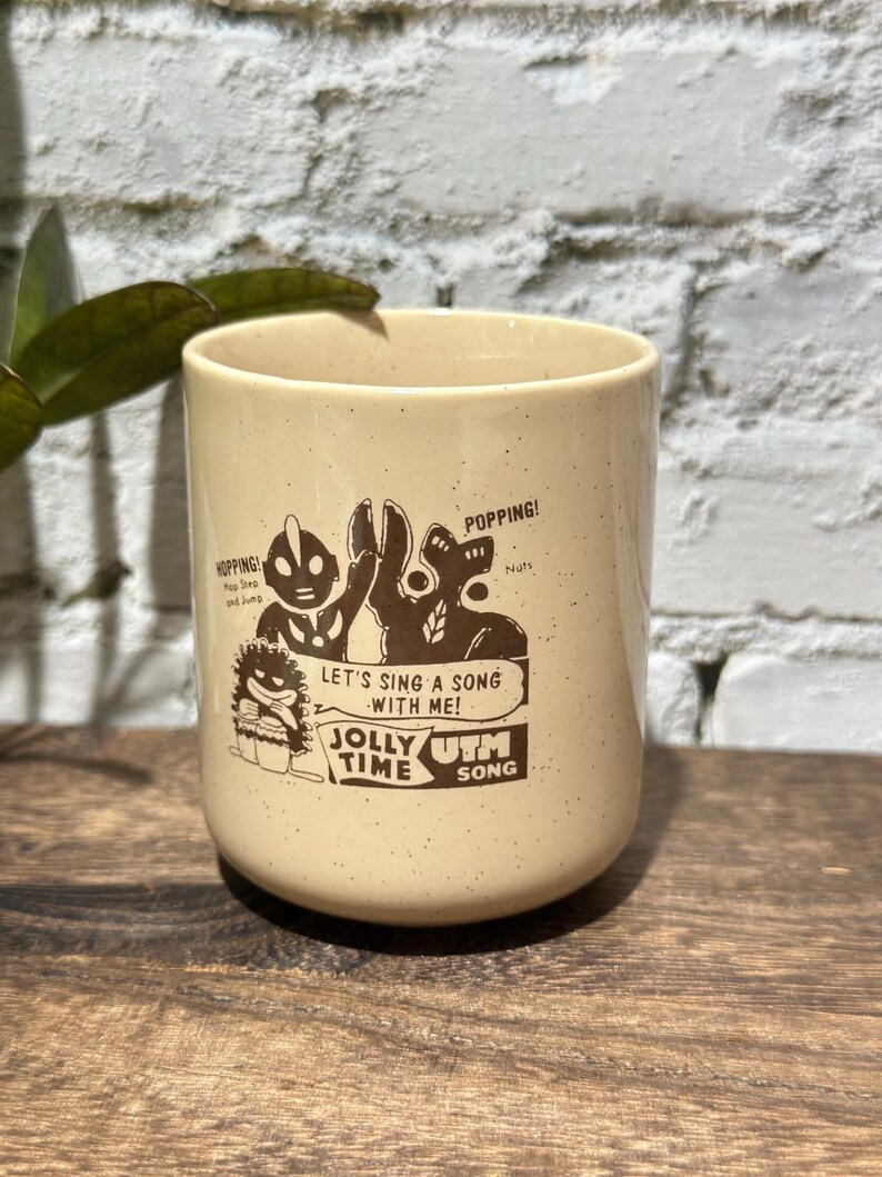 May include: A cream-colored ceramic mug with brown speckles featuring a graphic illustration of characters and the text "HOPPING! Hop Step and Jump", "POPPING! Nuts", "LET'S SING A SONG WITH ME!", "JOLLY TIME", and "UTM SONG" printed in brown ink.