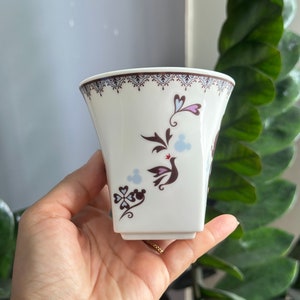 May include: A white ceramic cup with a brown and blue floral design featuring a bird and a Mickey Mouse silhouette. The cup has a square base and a scalloped rim.