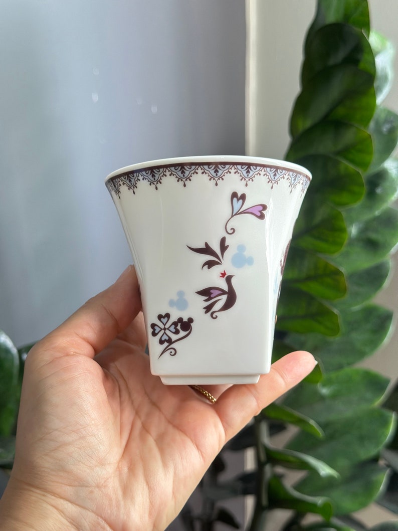 May include: A white ceramic cup with a brown and blue floral design featuring a bird and a Mickey Mouse silhouette. The cup has a square base and a scalloped rim.