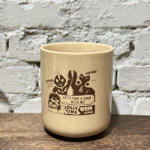 May include: A cream-colored ceramic mug with brown speckled details and a dark brown graphic print. The design features cartoon characters, the text "HOPPING!", "POPPING!", "LET'S SING A SONG WITH ME!", "JOLLY TIME", and "UTM SONG" printed on the front.