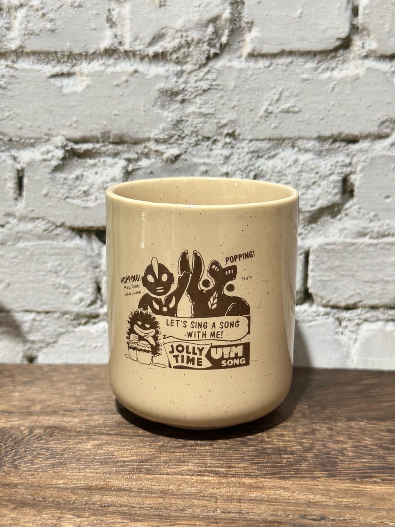May include: A cream-colored ceramic mug with brown speckled details and a dark brown graphic print. The design features cartoon characters, the text "HOPPING!", "POPPING!", "LET'S SING A SONG WITH ME!", "JOLLY TIME", and "UTM SONG" printed on the front.
