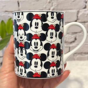 Puede incluir: Taza de cer&aacute;mica blanca con un patr&oacute;n repetido de caras de Mickey Mouse con sombreros rojos. La taza tiene un asa blanca y un borde negro. El dise&ntilde;o incluye varias expresiones de Mickey Mouse.