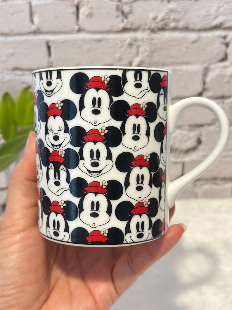 Puede incluir: Taza de cer&aacute;mica blanca con un patr&oacute;n repetido de caras de Mickey Mouse con sombreros rojos. La taza tiene un asa blanca y un borde negro. El dise&ntilde;o incluye varias expresiones de Mickey Mouse.