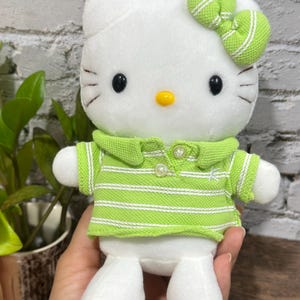 Pode incluir: Um brinquedo de pelúcia branco com olhos ovais pretos, nariz amarelo e três bigodes de cada lado. A personagem veste uma camisa polo verde-limão com gola, listras horizontais brancas finas e dois botões, além de um laço verde a combinar.