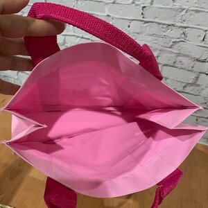 K&ouml;nnte beinhalten: Eine wiederverwendbare rosa Einkaufstasche mit zwei rosa Stoffgriffen. Die Tasche ist aus leichtem Material und hat einen flachen Boden.