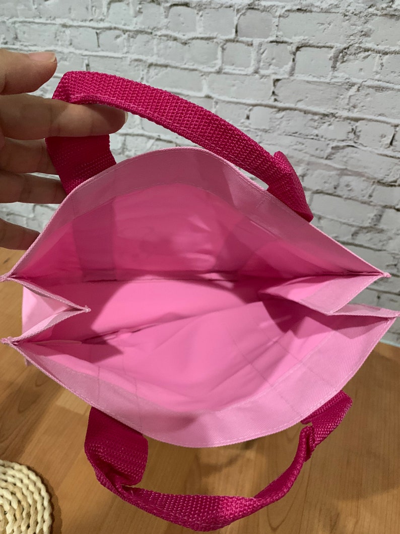 K&ouml;nnte beinhalten: Eine wiederverwendbare rosa Einkaufstasche mit zwei rosa Stoffgriffen. Die Tasche ist aus leichtem Material und hat einen flachen Boden.
