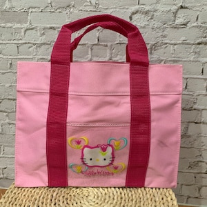 K&ouml;nnte beinhalten: Rosa Hello Kitty Tragetasche mit rosa Henkeln und einem Hello Kitty Design mit Blumen.