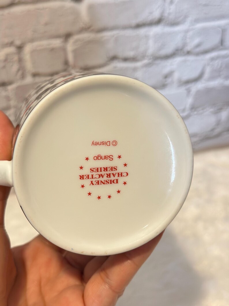 Puede incluir: Taza de cer&aacute;mica blanca con un patr&oacute;n en blanco y negro en el borde. La base de la taza presenta texto rojo que dice "DISNEY CHARACTER SERIES" y "Sango" con el logotipo de Disney.