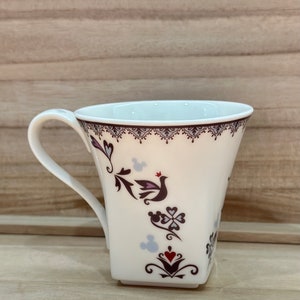May include: White ceramic mug with a square base and a handle. The mug features a floral design with a bird and Mickey Mouse silhouettes in shades of purple, red, and black.