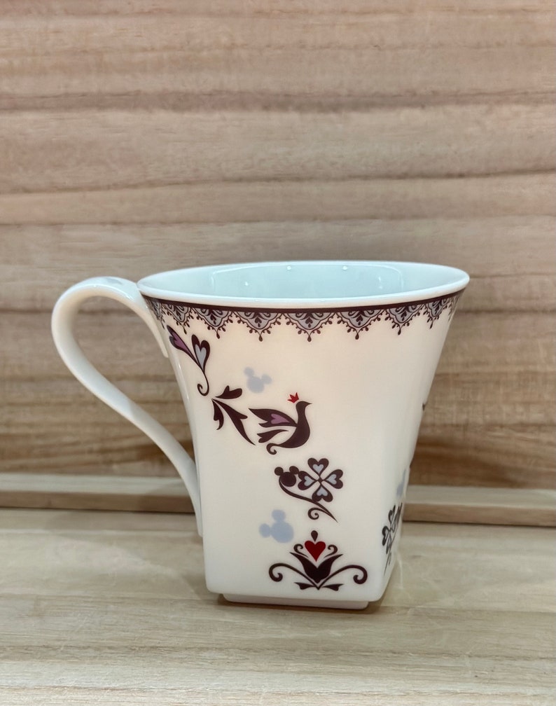 May include: White ceramic mug with a square base and a handle. The mug features a floral design with a bird and Mickey Mouse silhouettes in shades of purple, red, and black.