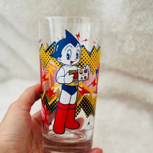 May include: A clear glass featuring a colourful cartoon illustration of Astro Boy, a Japanese manga character, holding a device. The glass has a red, yellow and black design with a white background.