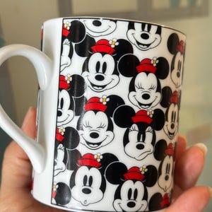 Puede incluir: Taza de cer&aacute;mica blanca con un patr&oacute;n repetido de caras de Mickey y Minnie Mouse. Los personajes de dibujos animados son en blanco, negro y rojo, con Minnie usando un sombrero rojo. La taza tiene un asa blanca.