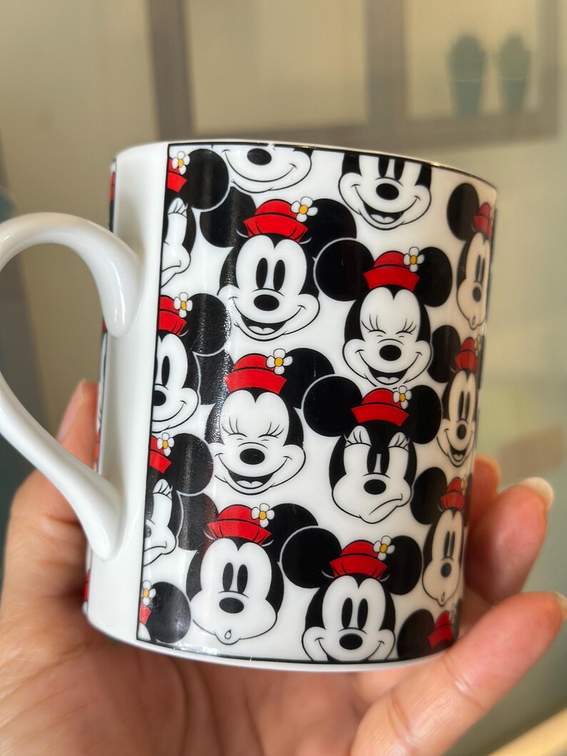 Puede incluir: Taza de cer&aacute;mica blanca con un patr&oacute;n repetido de caras de Mickey y Minnie Mouse. Los personajes de dibujos animados son en blanco, negro y rojo, con Minnie usando un sombrero rojo. La taza tiene un asa blanca.