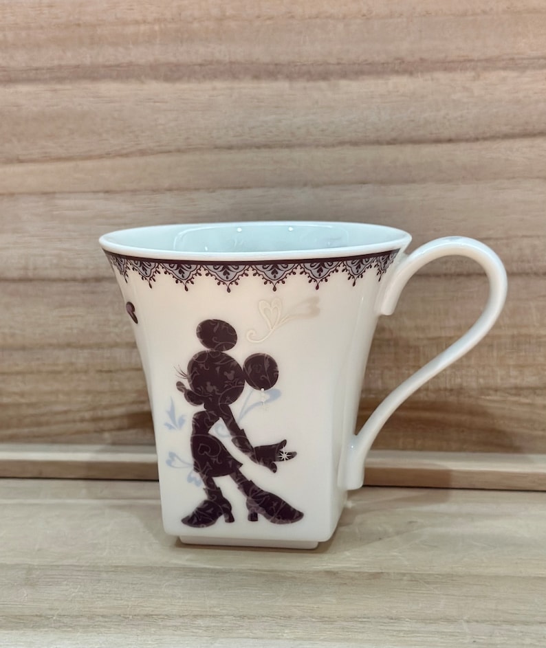 May include: White ceramic mug featuring a black silhouette of Minnie Mouse in a vintage-inspired design. The mug has a square base and a handle.