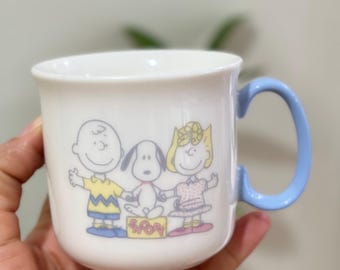 Può includere: Una tazza in ceramica bianca con un manico blu. La tazza presenta un'immagine di cartoni animati di Snoopy, Charlie Brown e Lucy della striscia a fumetti Peanuts. I personaggi sono in piedi su una scatola gialla con la parola "Snoopy" scritta su di essa.