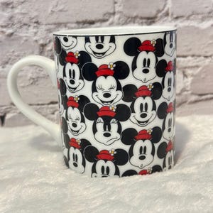 Puede incluir: Taza de cer&aacute;mica blanca con un patr&oacute;n repetido de caras de Mickey Mouse con sombreros rojos con flores amarillas. La taza tiene un asa blanca y un borde negro.