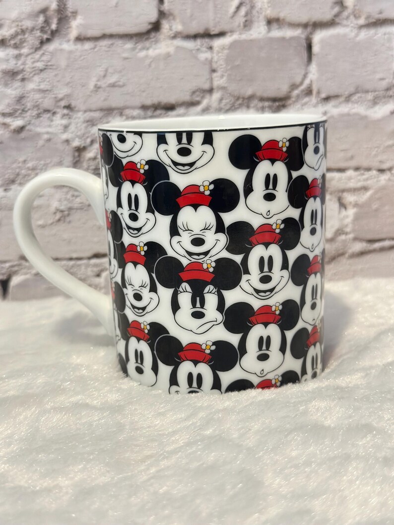 Puede incluir: Taza de cer&aacute;mica blanca con un patr&oacute;n repetido de caras de Mickey Mouse con sombreros rojos con flores amarillas. La taza tiene un asa blanca y un borde negro.