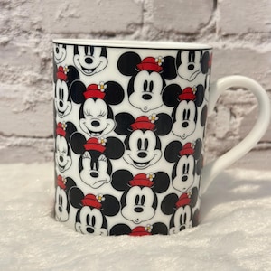 Puede incluir: Taza de cer&aacute;mica blanca con un patr&oacute;n repetido de caras de Mickey Mouse. Mickey lleva un sombrero rojo con una flor amarilla. La taza tiene un borde negro y un asa blanca.