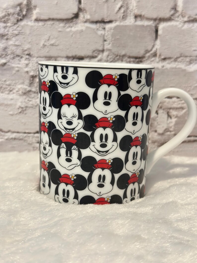 Puede incluir: Taza de cer&aacute;mica blanca con un patr&oacute;n repetido de caras de Mickey Mouse. Mickey lleva un sombrero rojo con una flor amarilla. La taza tiene un borde negro y un asa blanca.