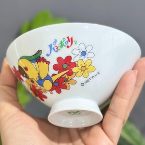 Pu&ograve; includere: Ciotola in ceramica bianca con un personaggio dei cartoni animati colorato e un design floreale. La ciotola presenta un personaggio giallo, rosso e blu con fiori e testo giapponese. La ciotola ha una piccola base.