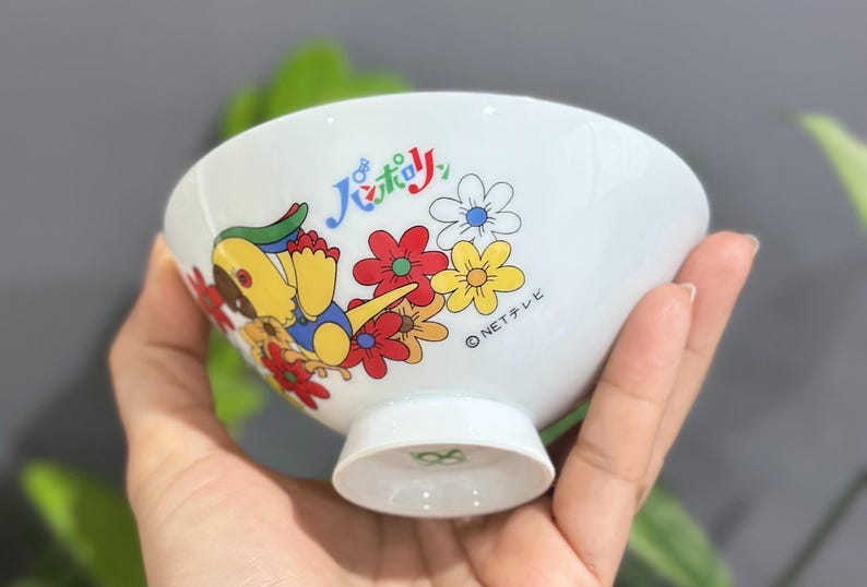 Pu&ograve; includere: Ciotola in ceramica bianca con un personaggio dei cartoni animati colorato e un design floreale. La ciotola presenta un personaggio giallo, rosso e blu con fiori e testo giapponese. La ciotola ha una piccola base.
