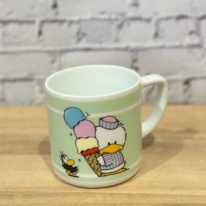 May include: A ceramic mug with a light green background and a cartoon illustration of a duck holding a large ice cream cone. The mug has a white handle and a white interior. A small black bird is also featured.