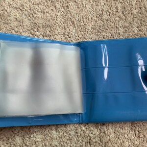 May include: A blue plastic wallet with clear plastic sleeves for holding cards or photos. The wallet has a snap closure and a small button on the side.
