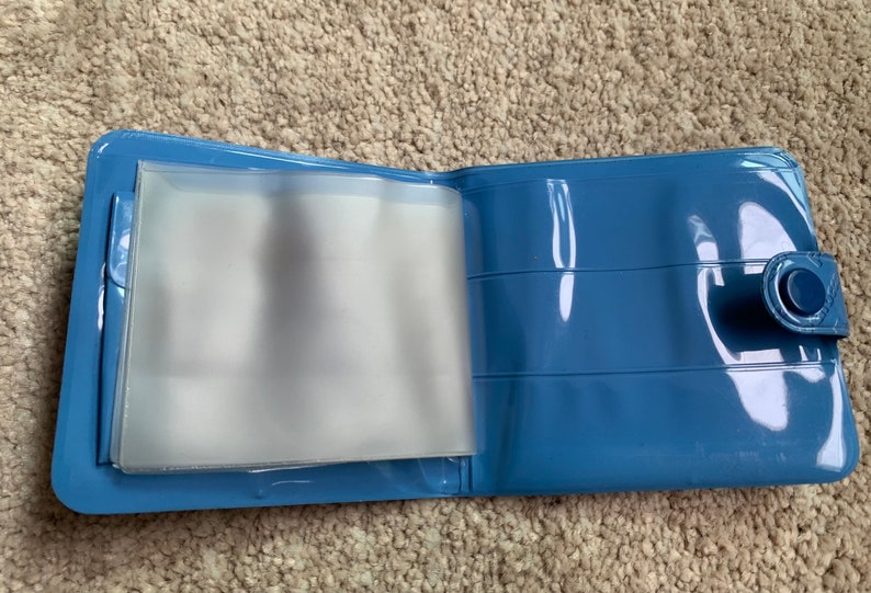 May include: A blue plastic wallet with clear plastic sleeves for holding cards or photos. The wallet has a snap closure and a small button on the side.