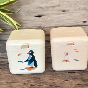 May include: Two off-white, square containers with hinged lids. Each container features a cartoon penguin illustration. One penguin is holding a pan, and the other is wearing a chef's hat. The containers are set on a wooden surface.