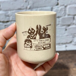 May include: A beige speckled ceramic mug featuring a dark brown graphic print of two characters, one resembling Ultraman, with the text "HOPPING! Hop Step and Jump", "POPPING! Nuts", "LET'S SING A SONG WITH ME!", and "JOLLY TIME UTM SONG".