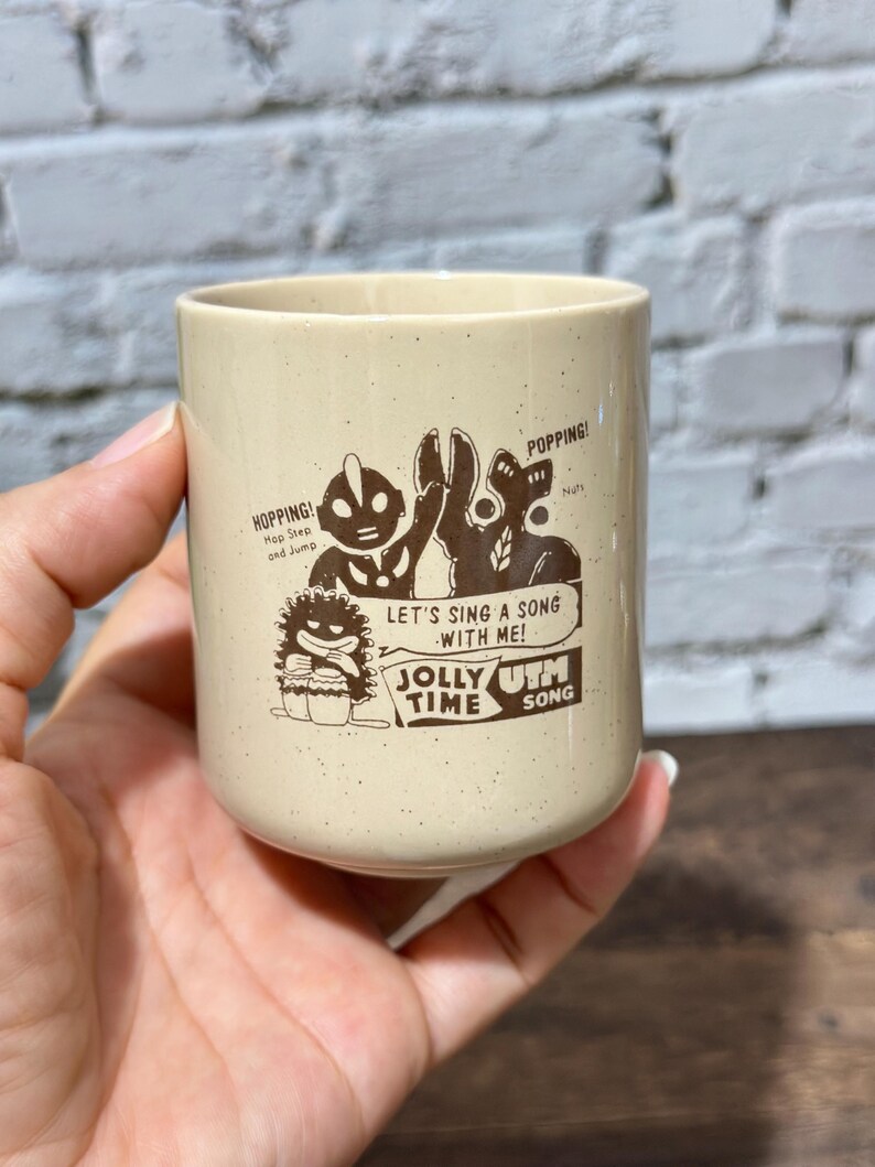 May include: A beige speckled ceramic mug featuring a dark brown graphic print of two characters, one resembling Ultraman, with the text "HOPPING! Hop Step and Jump", "POPPING! Nuts", "LET'S SING A SONG WITH ME!", and "JOLLY TIME UTM SONG".