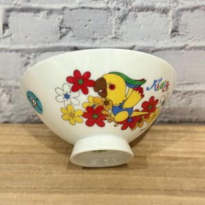 Pu&ograve; includere: Ciotola in ceramica bianca con un motivo di pappagallo e fiori colorati. Il pappagallo &egrave; giallo, blu e marrone, circondato da fiori rossi, gialli e bianchi. La ciotola ha una piccola base e testo in giapponese.