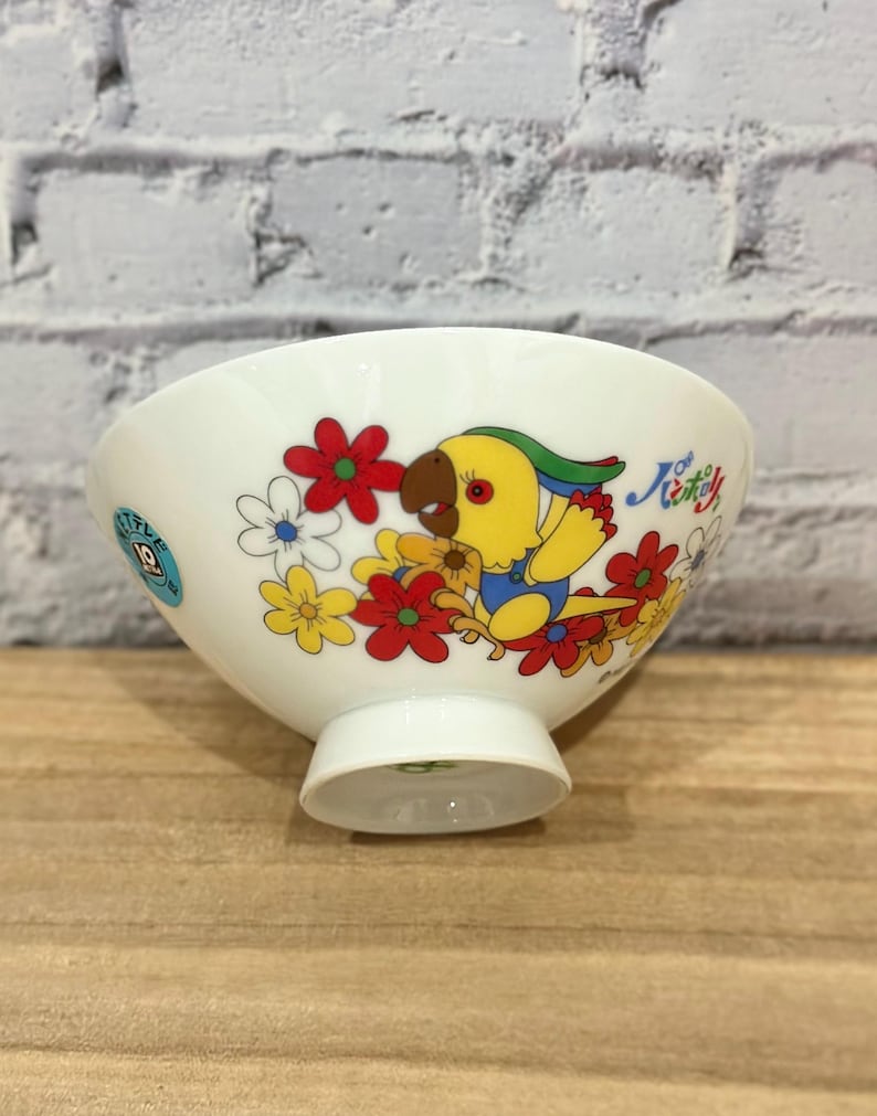 Pu&ograve; includere: Ciotola in ceramica bianca con un motivo di pappagallo e fiori colorati. Il pappagallo &egrave; giallo, blu e marrone, circondato da fiori rossi, gialli e bianchi. La ciotola ha una piccola base e testo in giapponese.