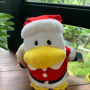 May include: A plush toy duck wearing a Santa hat and a red and white Santa suit. The duck is holding a brown sack in its left wing.