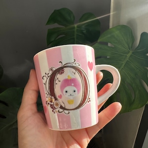May include: A pink and white striped ceramic mug featuring a cartoon character in a frame. The character is wearing a pink crown and holding a yellow cupcake. The mug has a brown handle.
