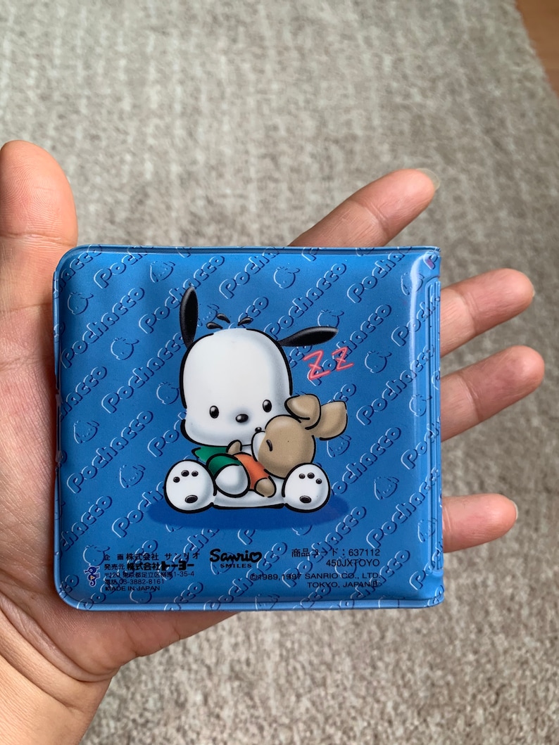 May include: A blue plastic coin purse with a white cartoon dog and a brown bunny. The purse has a repeating pattern of the dog's name "Pochacco" on the front and back. The purse has the text "Sanrio Smiles" and "450JXTOYO" on the bottom.