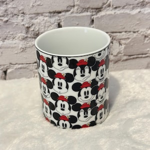Puede incluir: Una taza de cer&aacute;mica blanca con un patr&oacute;n repetido de la cara de Minnie Mouse. Minnie se representa con orejas negras, un sombrero rojo y una expresi&oacute;n sonriente. La taza tiene un interior blanco y un borde negro.