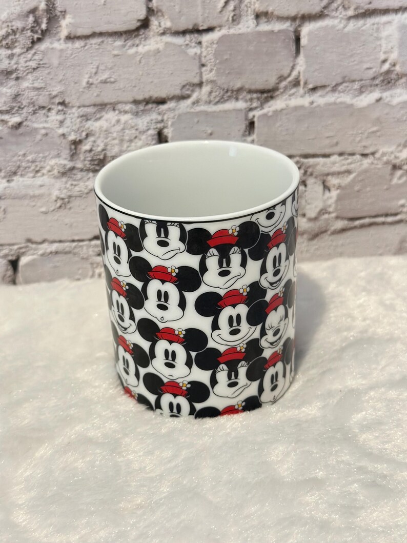 Puede incluir: Una taza de cer&aacute;mica blanca con un patr&oacute;n repetido de la cara de Minnie Mouse. Minnie se representa con orejas negras, un sombrero rojo y una expresi&oacute;n sonriente. La taza tiene un interior blanco y un borde negro.