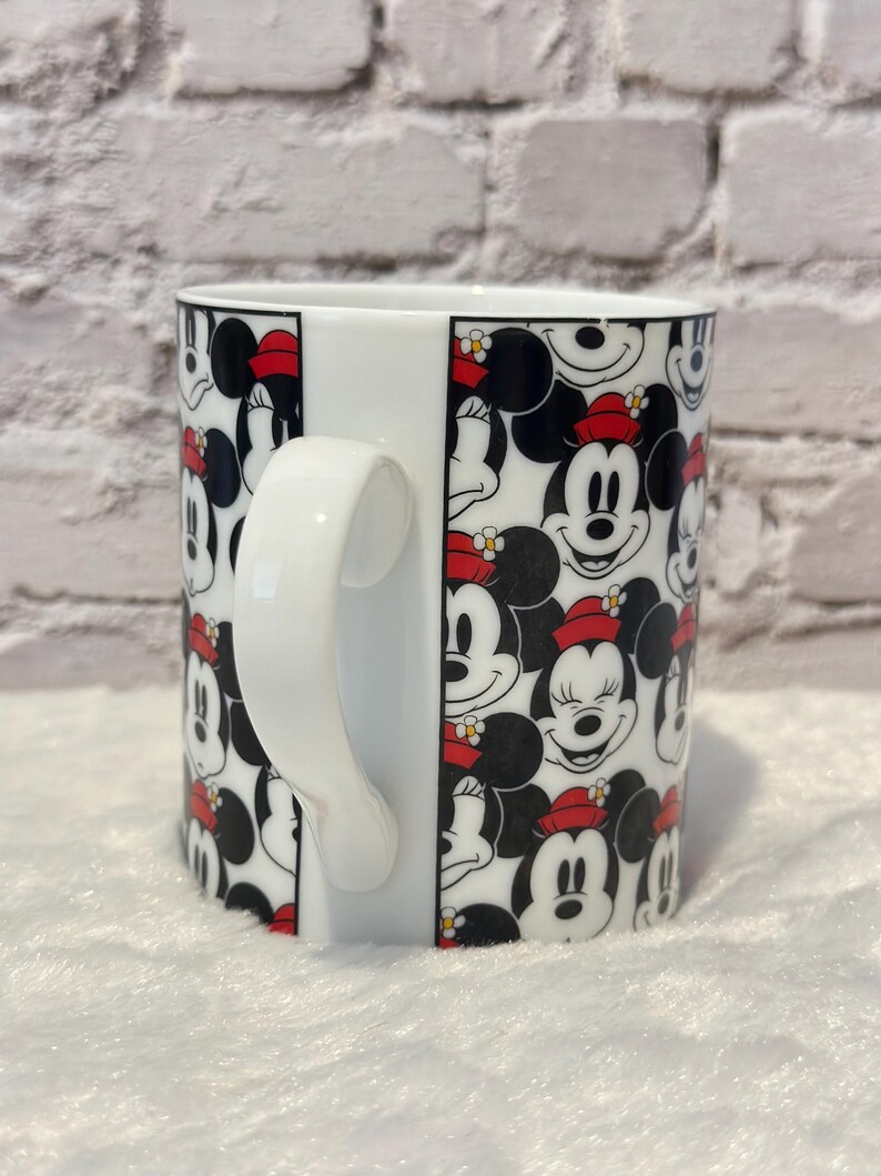 Puede incluir: Taza de cer&aacute;mica blanca con un patr&oacute;n repetido de caras de Mickey y Minnie Mouse. Los personajes son en blanco, negro y rojo, con Minnie usando un sombrero rojo. La taza tiene un asa blanca y un acabado brillante.