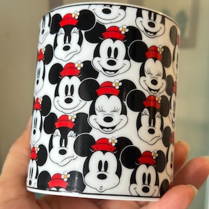 Puede incluir: Una taza cil&iacute;ndrica de cer&aacute;mica con un patr&oacute;n repetido de caras de Mickey y Minnie Mouse. Las caras en blanco y negro est&aacute;n adornadas con sombreros rojos y detalles florales, sobre un fondo blanco.