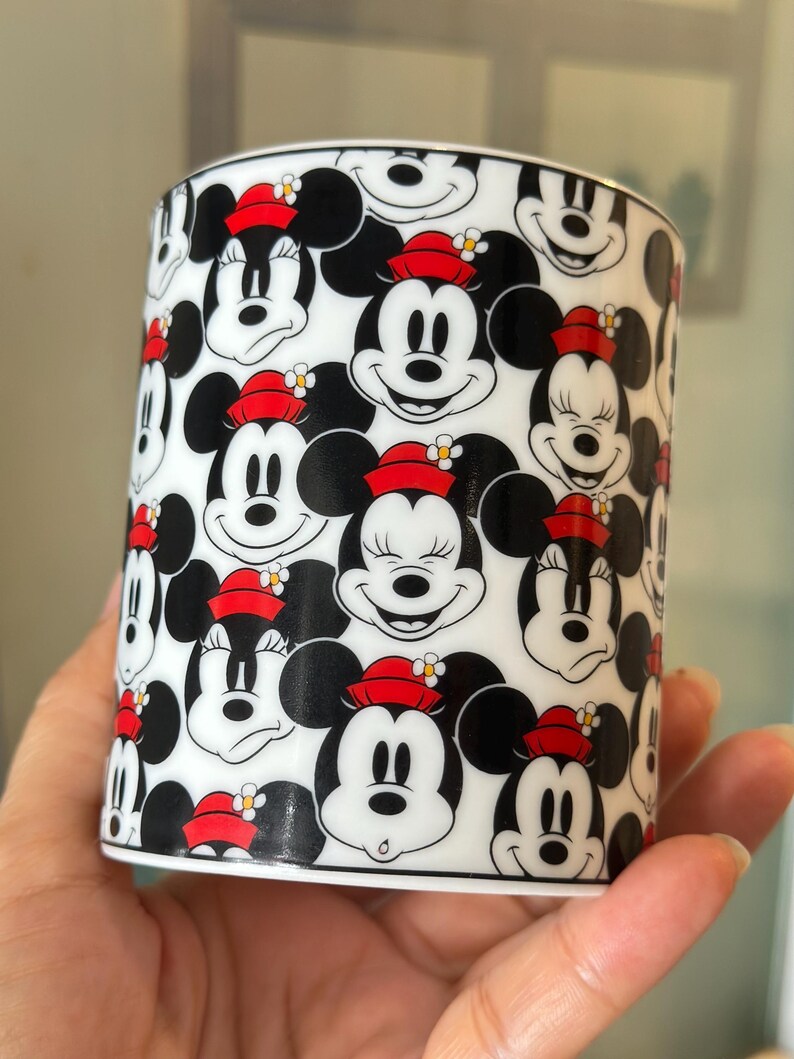 Puede incluir: Una taza cil&iacute;ndrica de cer&aacute;mica con un patr&oacute;n repetido de caras de Mickey y Minnie Mouse. Las caras en blanco y negro est&aacute;n adornadas con sombreros rojos y detalles florales, sobre un fondo blanco.