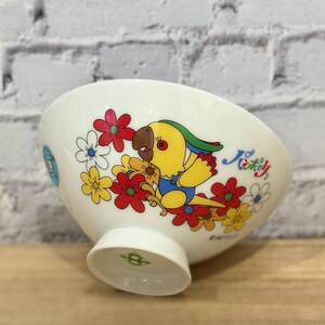 Pu&ograve; includere: Ciotola in ceramica bianca con un colorato design di pappagallo e fiori. Il pappagallo &egrave; giallo con testa verde e tuta blu, circondato da fiori rossi, gialli e bianchi. La ciotola ha un piedino e un disegno decorativo sul lato.
