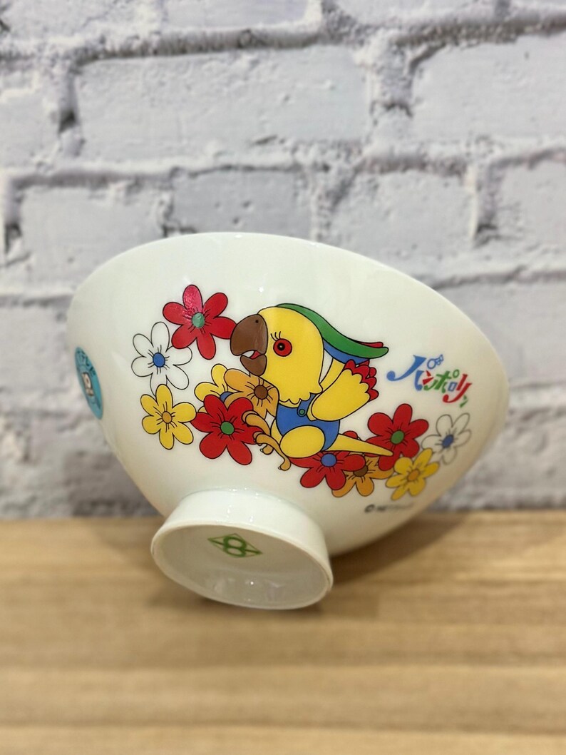 Pu&ograve; includere: Ciotola in ceramica bianca con un colorato design di pappagallo e fiori. Il pappagallo &egrave; giallo con testa verde e tuta blu, circondato da fiori rossi, gialli e bianchi. La ciotola ha un piedino e un disegno decorativo sul lato.
