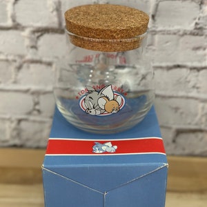 May include: A glass jar with a cork lid featuring Tom and Jerry cartoon characters. The jar is in a blue box with a red stripe and a Tom and Jerry cartoon character on the front.