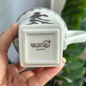 May include: White ceramic mug featuring a brown silhouette of Mickey Mouse and Minnie Mouse on the front. The base of the mug displays the text "DRAMATIC DisneySEA 2005 &copy;DISNEY".