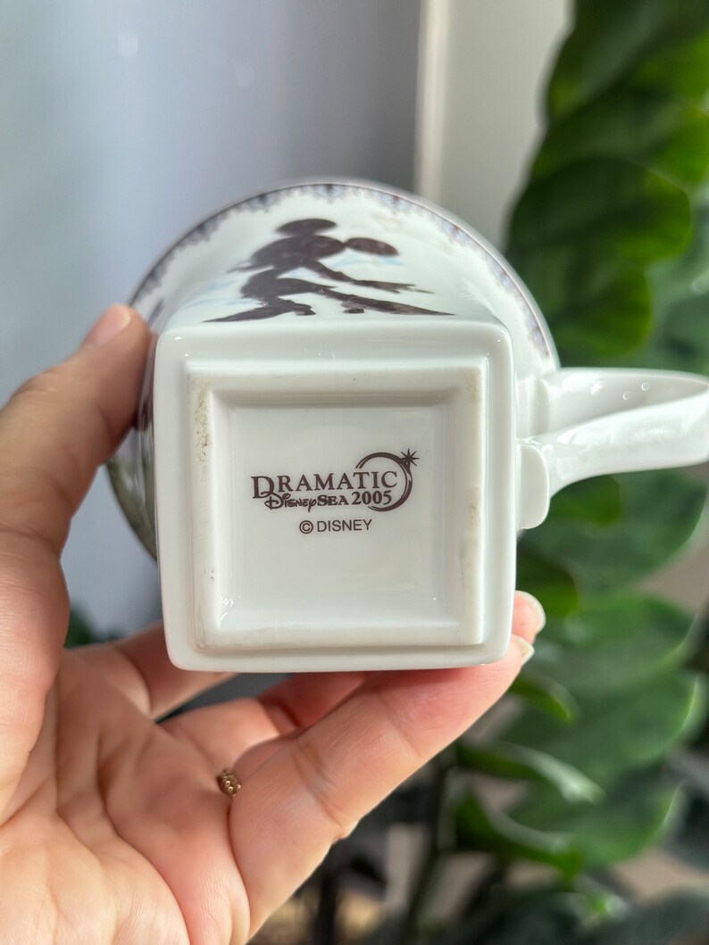 May include: White ceramic mug featuring a brown silhouette of Mickey Mouse and Minnie Mouse on the front. The base of the mug displays the text "DRAMATIC DisneySEA 2005 &copy;DISNEY".